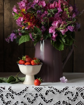 Bouquet di fiori da giardino estivo e fragole sul tavolo con una tovaglia di pizzo bianco interno rustico phlox snapdragon ageratum in una brocca