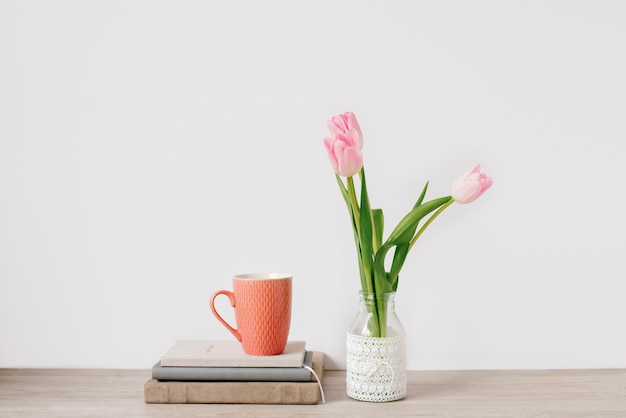 Bouquet of three pink tulips in a glass vase and a orange cup on top of books on white background