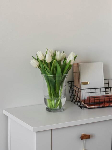 A bouquet of tulips in a glass vase a metal basket with notebooks books on a white wooden chest of drawers Minimalism style