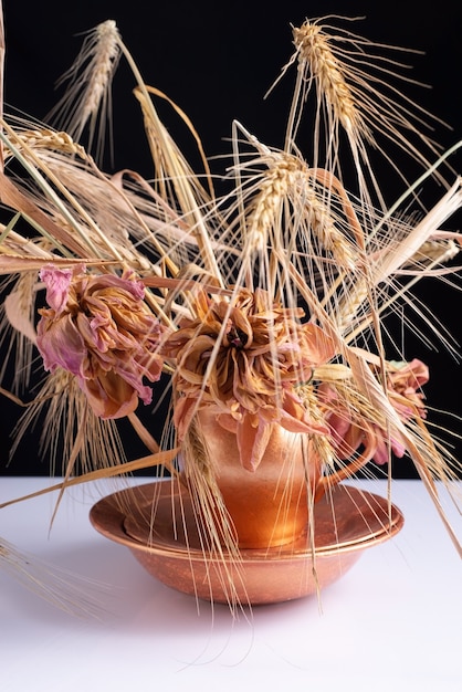 Un mazzo di spighe di grano con fiori secchi in un piatto di rame su un tavolo bianco e uno sfondo nero.