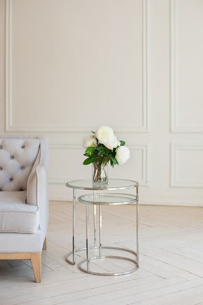 Bouquet of white peonies stands in vase on table near gray sofa in white living room.