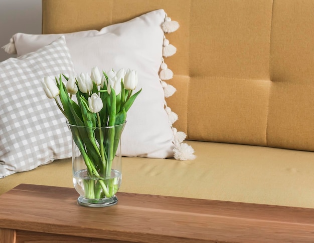 A bouquet of white tulips in a glass vase on a wooden table next to a yellow sofa in the living room
