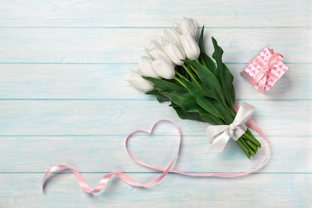 A bouquet of white tulips and a pink ribbon in the form of a heart with a gift box on blue wooden boards . 