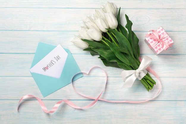 白いチューリップの花束とギフトボックス、ハートの形をしたハート型のピンクのリボン、青い木の板に封筒。母の日