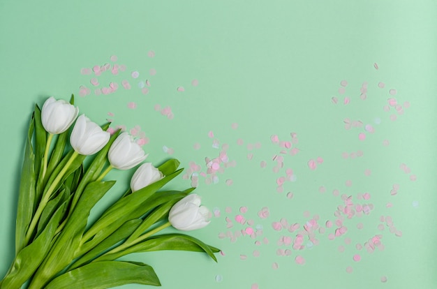 Bouquet of white tulips with colored confetti on a green table with copy space. Spring concept, flat lay, place for text. View from above.