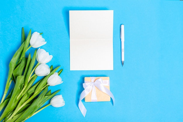 Bouquet of white tulips with gift boxes and blank paper on a blue surface