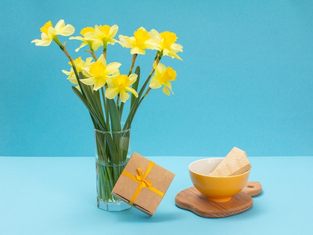 Bouquet of yellow daffodils in glass vase a gift box and a plate with waffles on a blue background