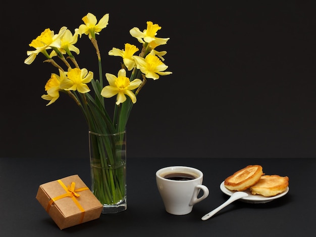 Bouquet of yellow daffodils in a vase gift box a cup of coffee and a plate with fritters and spoon