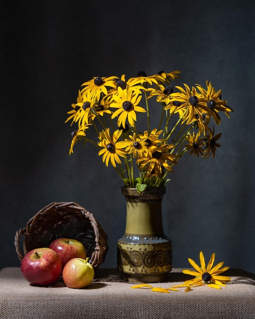 A bouquet of yellow daisies in a ceramic vase and three red apples in a basket on the table Grey background Still life