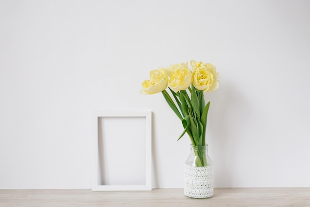 A bouquet of yellow spring tulips in a vase stands on the table next to an empty white frame for a photo with copy space