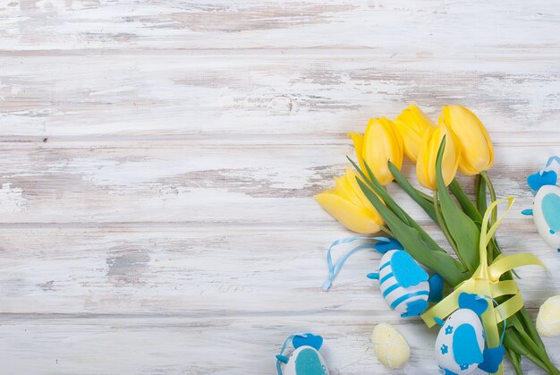 Bouquet of yellow tulips and chikken easter eggs with a blue ribbon on a wooden background