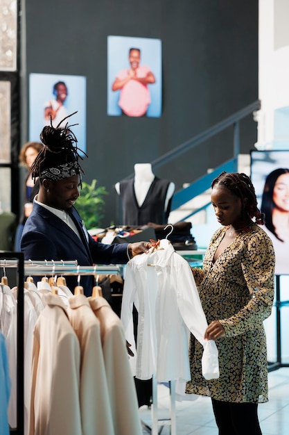 사진 부티크 직원은 고객에게 캐주얼한 옷을 입히고 옷가게에서 직물에 대해 논의하는 흰색 셔츠를 보고 있습니다. 출산 옷과 유행 상품을 구입하는 아프리카 계 미국인 고객