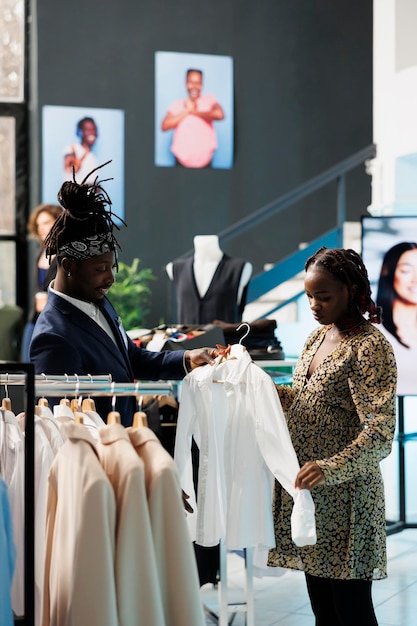 Boutique-medewerker helpt klant met vrijetijdskleding, kijkend naar wit overhemd dat stof bespreekt in kledingwinkel. Afro-Amerikaanse klant die zwangerschapskleding en trendy merchandise koopt