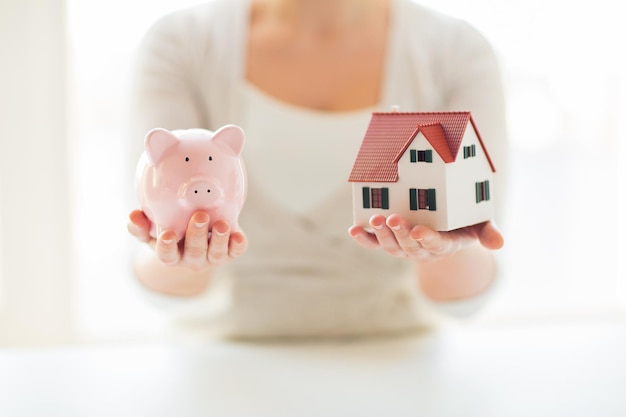 Bouw-, hypotheek-, investerings-, onroerend goed- en eigendomsconcept - close-up van vrouw met huis- of huismodel en spaarvarken