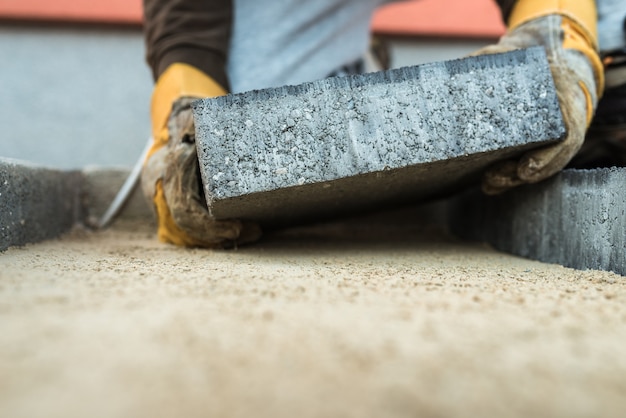 Bouwer die een straatsteen legt en deze met gehandschoende handen op de zandfundering plaatst