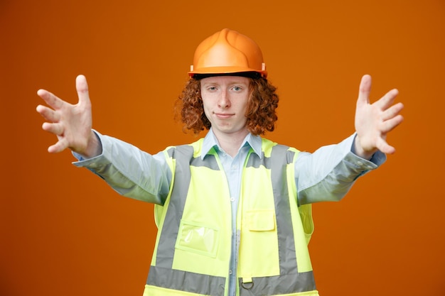 Bouwer jonge man in bouw uniform en veiligheidshelm kijken camera gelukkig en positief wijd openende handen groet gebaar staande over oranje achtergrond