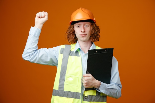 Bouwer jonge man in bouw uniform en veiligheidshelm met klembord met gebalde vuist kijkend naar camera met zelfverzekerde uitdrukking staande over oranje achtergrond