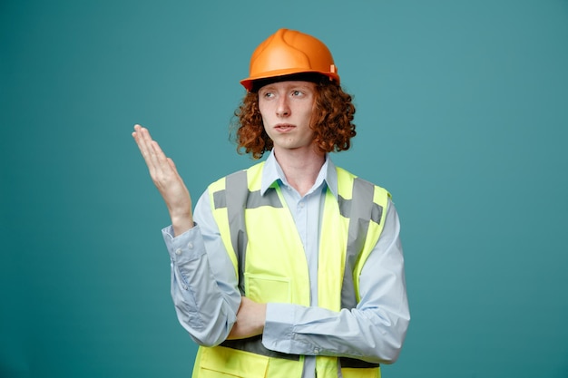 Bouwer jonge man in bouw uniform en veiligheidshelm op zoek opzij verward en ontevreden verhogen arm in ongenoegen staande over blauwe achtergrond