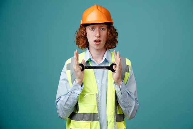 Bouwer jonge man in bouwuniform en veiligheidshelm met moersleutel kijkend naar camera met serieuze zelfverzekerde uitdrukking over blauwe achtergrond