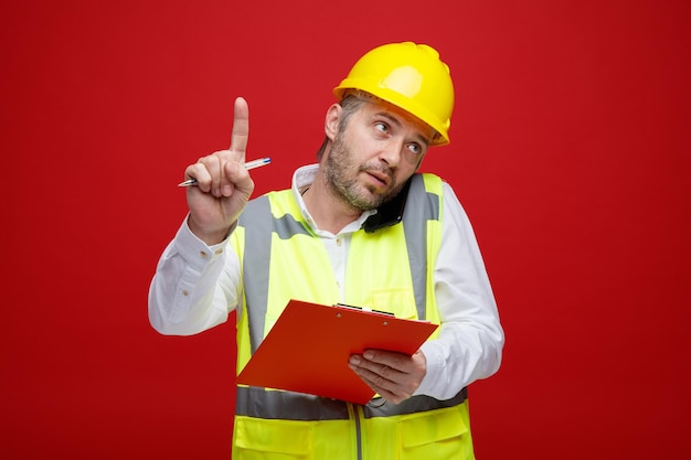 Bouwer man in bouw uniform en veiligheidshelm met klembord praten op mobiele telefoon ziet er zelfverzekerd uit en vraagt om te wachten met wijsvinger die over rode achtergrond staat