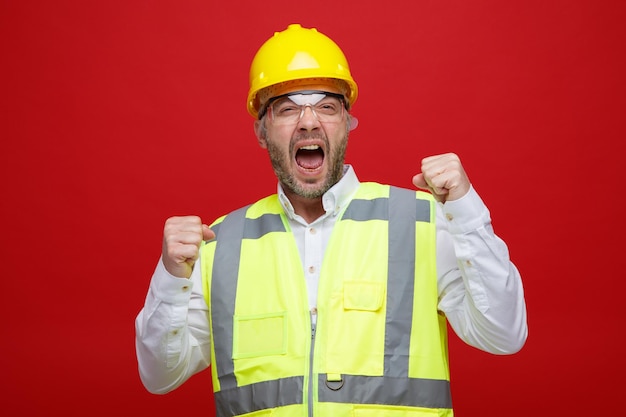 Bouwer man in bouw uniform en veiligheidshelm met veiligheidsbril boos en gefrustreerd gebalde vuisten schreeuwend over rode achtergrond