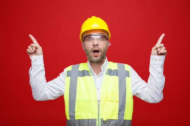 Bouwer man in bouw uniform en veiligheidshelm met veiligheidsbril kijkend naar camera verrast en verbaasd wijzend met wijsvingers omhoog staande over roze achtergrond