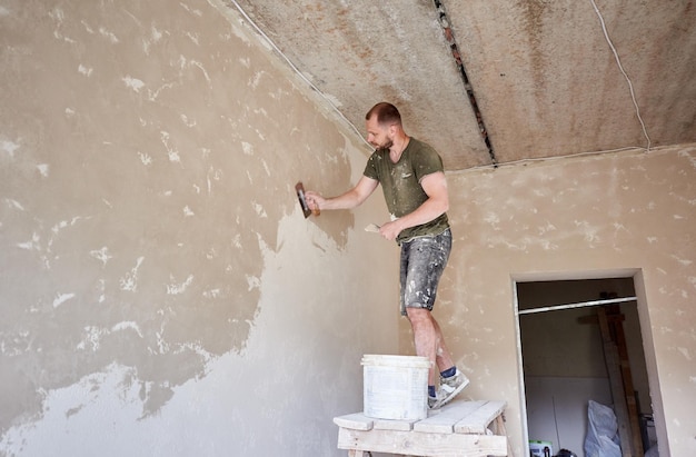 Bouwer staat op houten standaard en werkt met spatel met gips aan de muur in kleine kamer