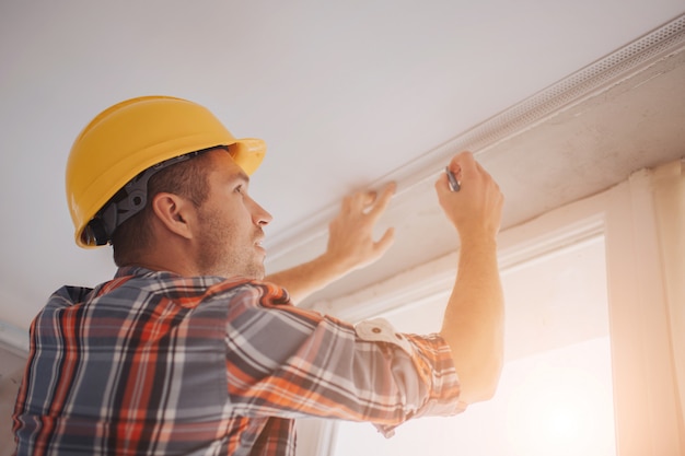 bouwer werkt op de bouwplaats en meet het plafond. Werknemer in een oranje bouwhelm maakt reparaties in huis.
