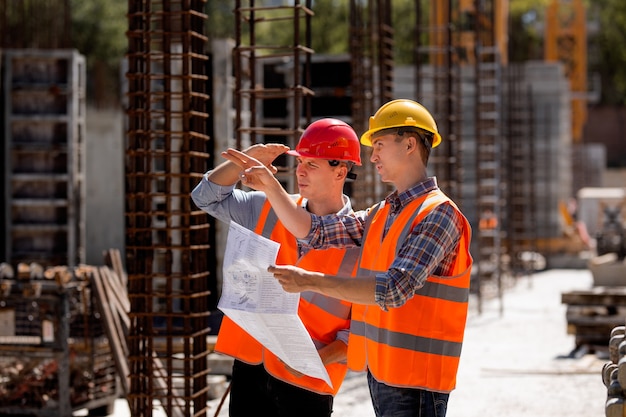 Bouwkundig ingenieur en bouwmanager in oranje werkvesten en harde helmen verkennen bouwdocumentatie op de bouwplaats bij de stalen frames. .