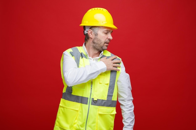 Bouwman in bouwuniform in veiligheidshelm die er onwel uitziet en zijn schouder aanraakt, voelt pijn die over rode achtergrond staat