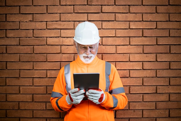 Bouwvakker in veiligheidsuitrusting die tegen bakstenen muur staat en tabletcomputer vasthoudt