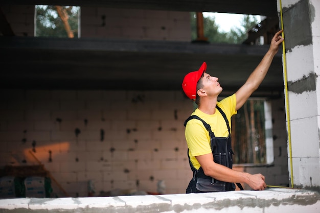 Bouwvakker op bouwplaats meet de lengte van raamopening en bakstenen muur met meetlint Cottage zijn gemaakt van poreuze betonblokken werkkleding jumpsuit en baseballcap