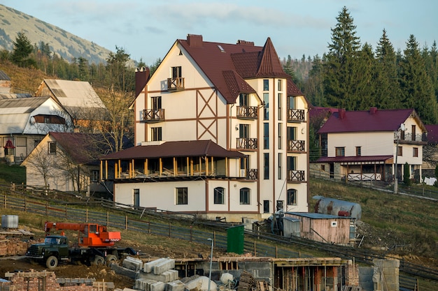 Bouwwerf met de bouw van kraan en nieuw modern comfortabel hotelhuis met dakspanendak op ecologisch plattelandsgebied op nette bomen en bergenheuvels onder blauwe hemel.