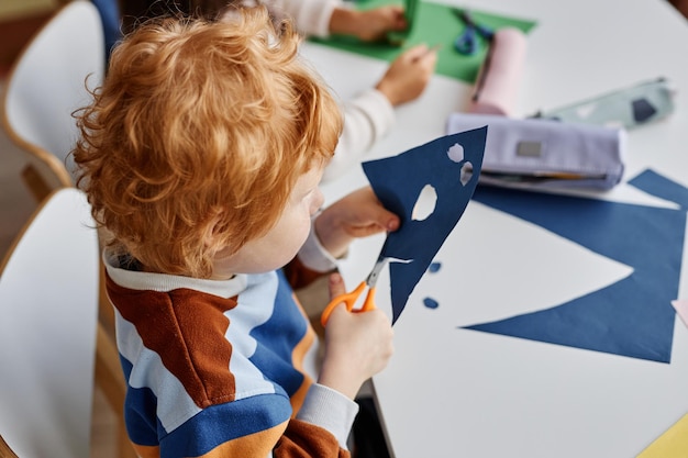 Boven de hoek van een schattige ijverige schooljongen die gaten in donkerblauw papier snijdt terwijl hij een masker maakt bij lesso