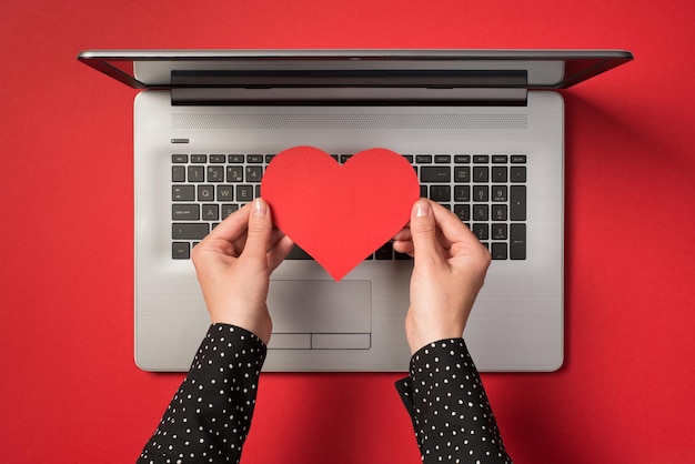 Boven foto van grijze laptop en handen met een rood papieren hart geïsoleerd op de rode achtergrond