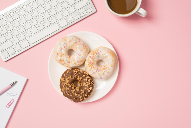 Boven foto van toetsenbord kopje koffie notebook met pen paperclips en plaat van geglazuurde donuts geïsoleerd op de roze achtergrond met lege ruimte