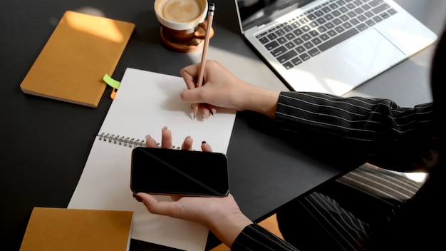 Boven geschoten van onderneemster die op notitieboekje schrijven terwijl het gebruiken van smartphone in comfortabele werkplaats