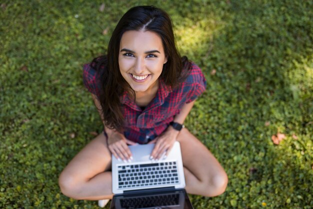 Boven het zicht van een jonge blanke brunette vrouw die lacht en een geruit hemd draagt, zittend op het gras voor een laptop die werkt op haar notebookcomputer met draadloze communicatie, winkelen en studeren