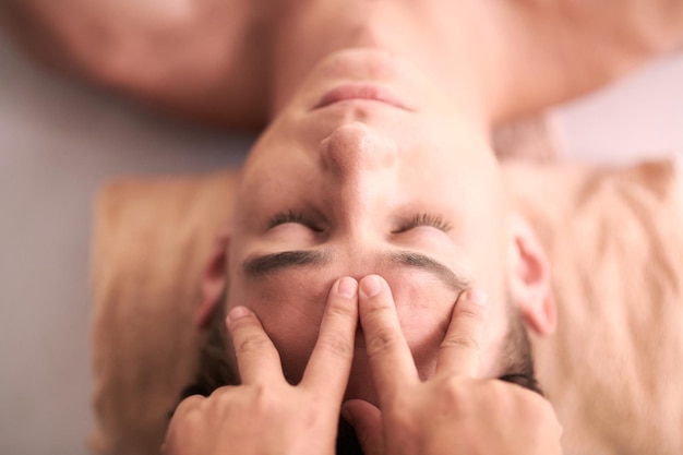 Foto boven hoek van jonge serene man die gezichtsmassage krijgt in een luxe spa salon terwijl masseuse zijn voorhoofd drukt tijdens acupressuur