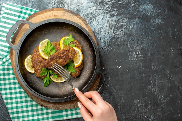 Boven weergave van hand met vork op heerlijke schnitzels geserveerd met greens en citroen in zwarte pan op houten bord op groene gestripte handdoek op donkere kleur achtergrond