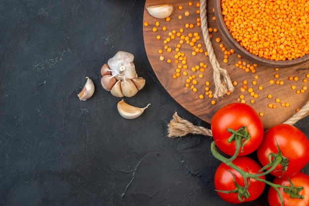 Boven weergave van rode linzen in een bruine kom touw knoflook op houten ronde bord tomaten met stengels op zwarte achtergrond