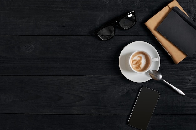 Boven weergave van slimme telefoon met notebook en kopje latte koffie op zwarte houten achtergrond