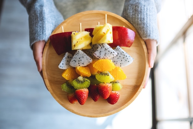 Foto bovenaanzicht afbeelding van een vrouw met een houten bord met vers gemengd fruit op spiesjes