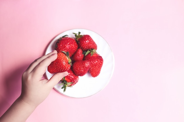 Bovenaanzicht Childs hand met een aardbei op roze tafel, plaat van aardbeien. Zomer gezond eten concept.