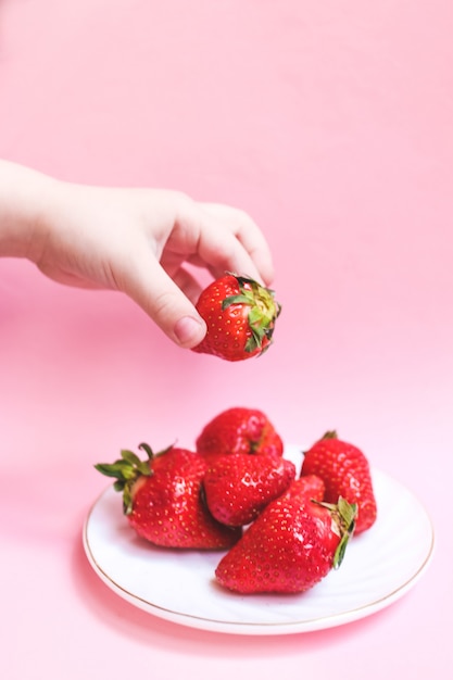 Foto bovenaanzicht childs hand met een aardbei op roze tafel, plaat van aardbeien. zomer gezond eten concept.