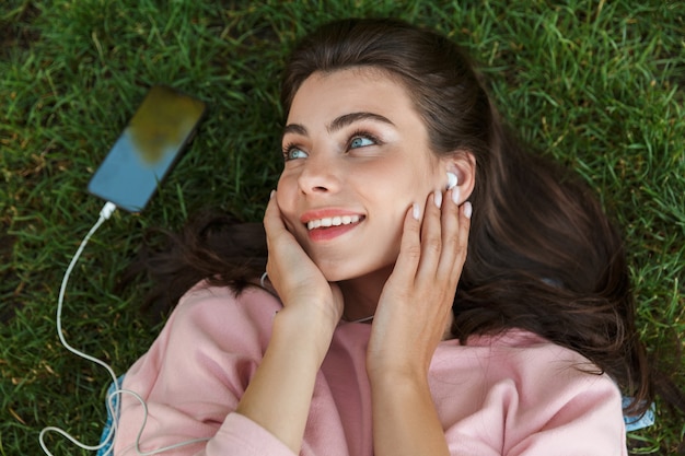 Foto bovenaanzicht close-up van een mooie jonge brunette vrouw die buiten op gras ligt, luisterend naar muziek met oortelefoons en mobiele telefoon met leeg scherm