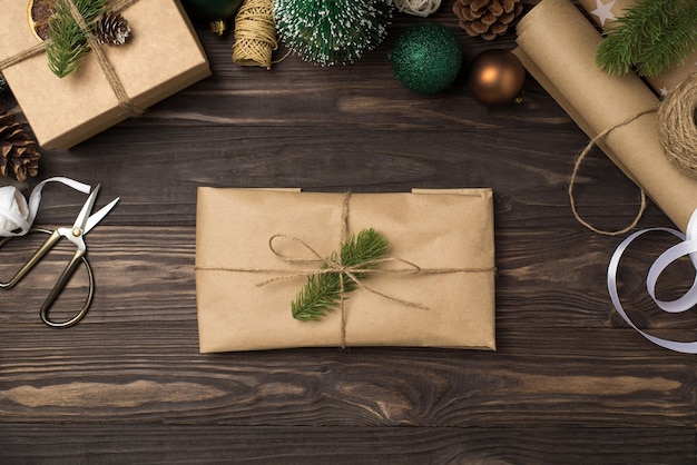 Bovenaanzicht foto van ambachtelijke papieren pakket versierd met grenen takje en touw boog handwerk tools kegels giftbox groene en gouden kerstboom ballen op geïsoleerde donkere houten bureau achtergrond
