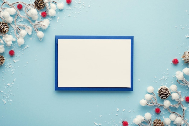 Bovenaanzicht foto van blauwe envelop met witte kaart kerstversiering sneeuw takken rode bessen en dennenappels op geïsoleerde pastel blauwe achtergrond met lege ruimte