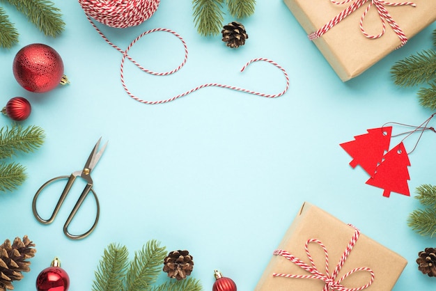 Bovenaanzicht foto van dennen twijgen kegels rode kerstboom ballen schaar spoel van decoratieve touw spar vormige tags en twee ambachtelijke papier geschenkdozen op geïsoleerde pastel blauwe achtergrond met copyspace