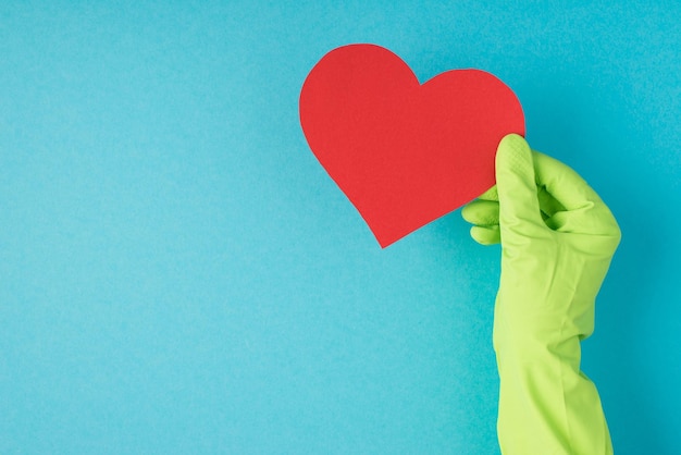 Bovenaanzicht foto van hand in groene rubberen handschoen met rood papieren hart op geïsoleerde pastelblauwe achtergrond met lege ruimte